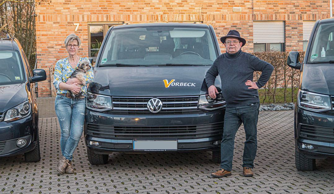 Zwei Personen stehen links und rechts neben einem PKW - erste Person hat einen Hund in den Armen, zweite Person trägt einen Hut